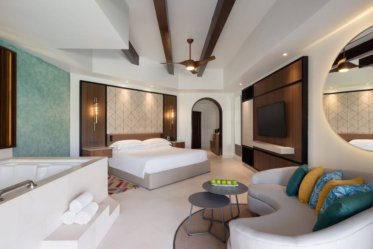 Una habitación de hotel moderna con una cama grande, un sofá, una mesita con manzanas y un televisor.