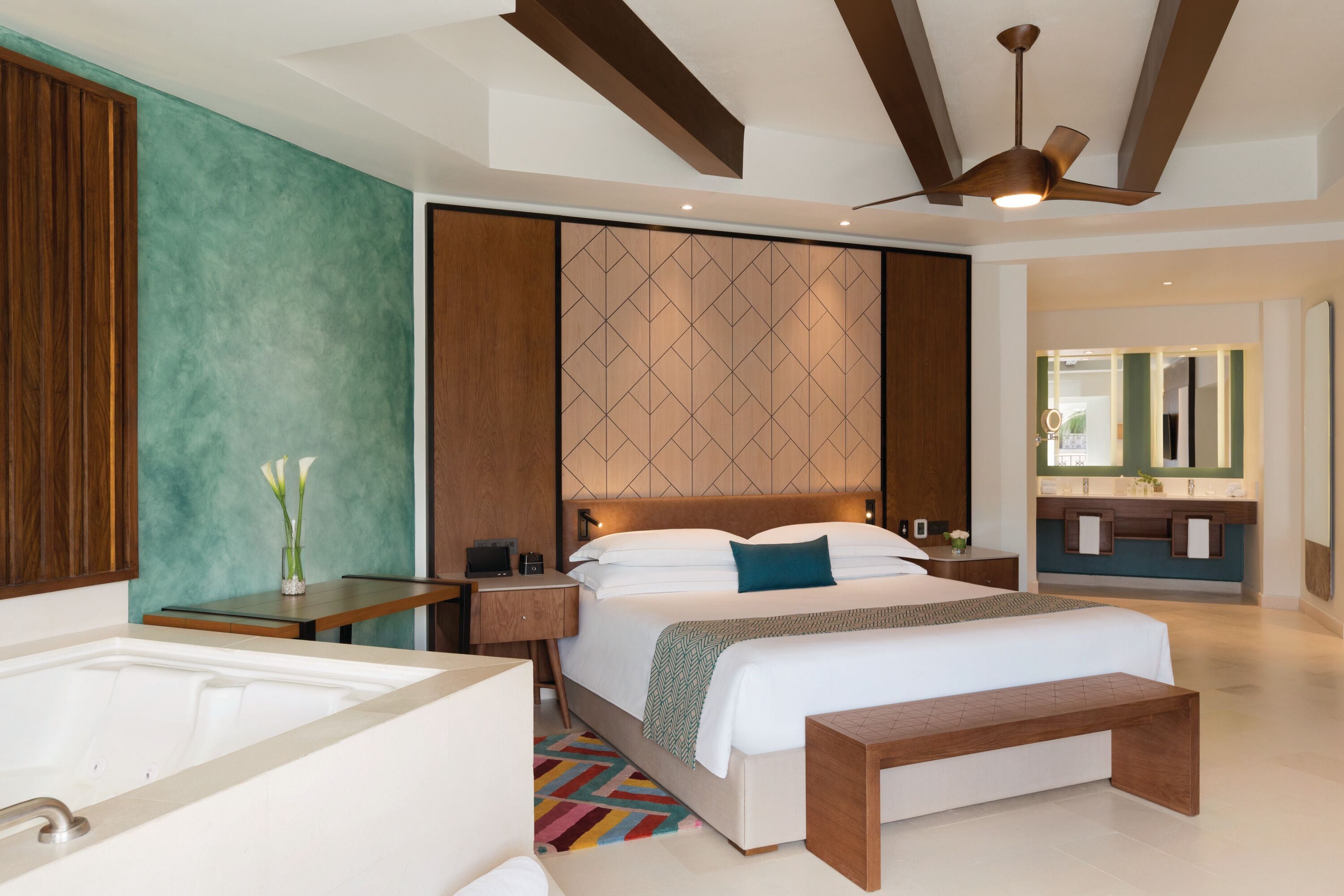 A modern hotel room with a large bed, a bedside table, a nightstand, a desk, a chair, a bench, a bathtub, and a ceiling fan.