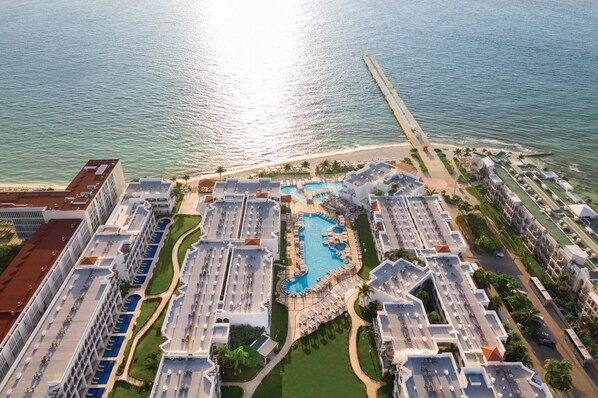 Vista aérea de un complejo turístico con varias edificaciones, una piscina central y una zona de playa.