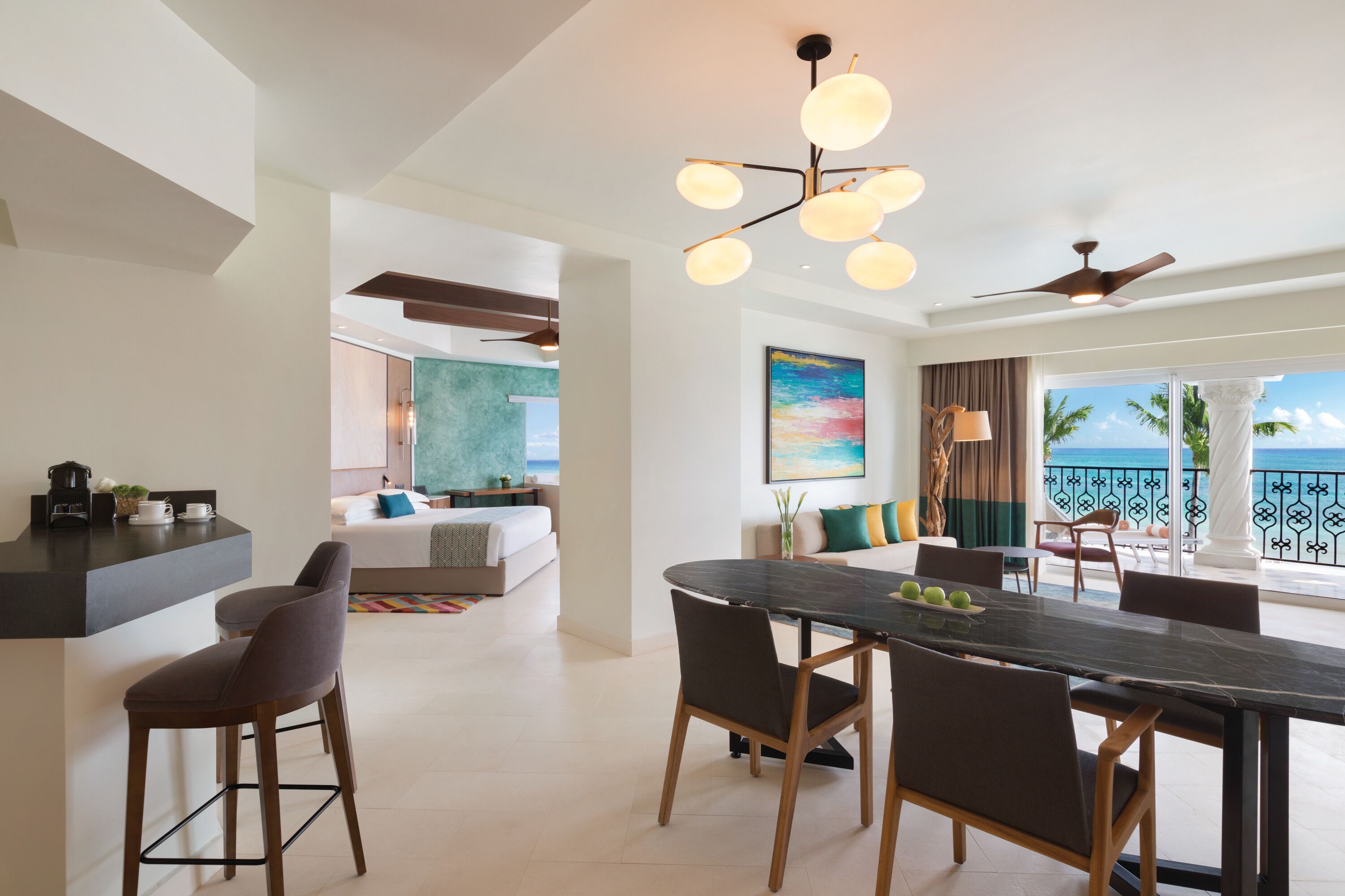 A modern hotel room with a dining area, a kitchen, and a balcony with a view of the sea.