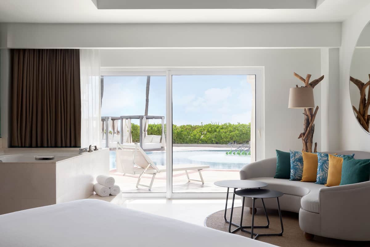 A modern hotel room with a large bed, a sofa, a small table, and a view of a pool and greenery.