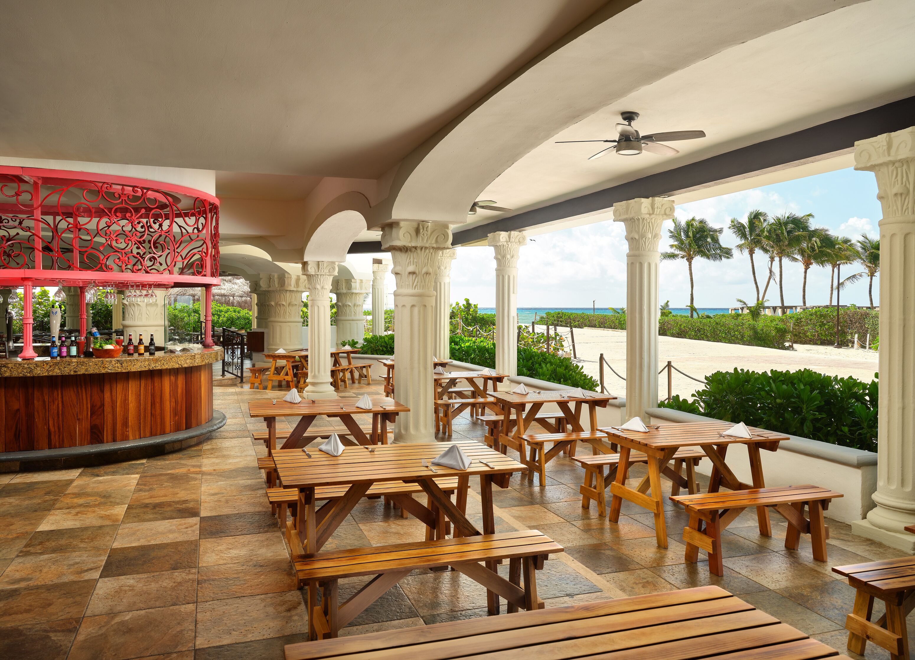 A covered outdoor dining area with wooden tables and benches, white columns, and a view of the beach.
