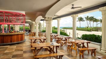 A covered outdoor dining area with wooden tables and benches, white columns, and a view of the beach.