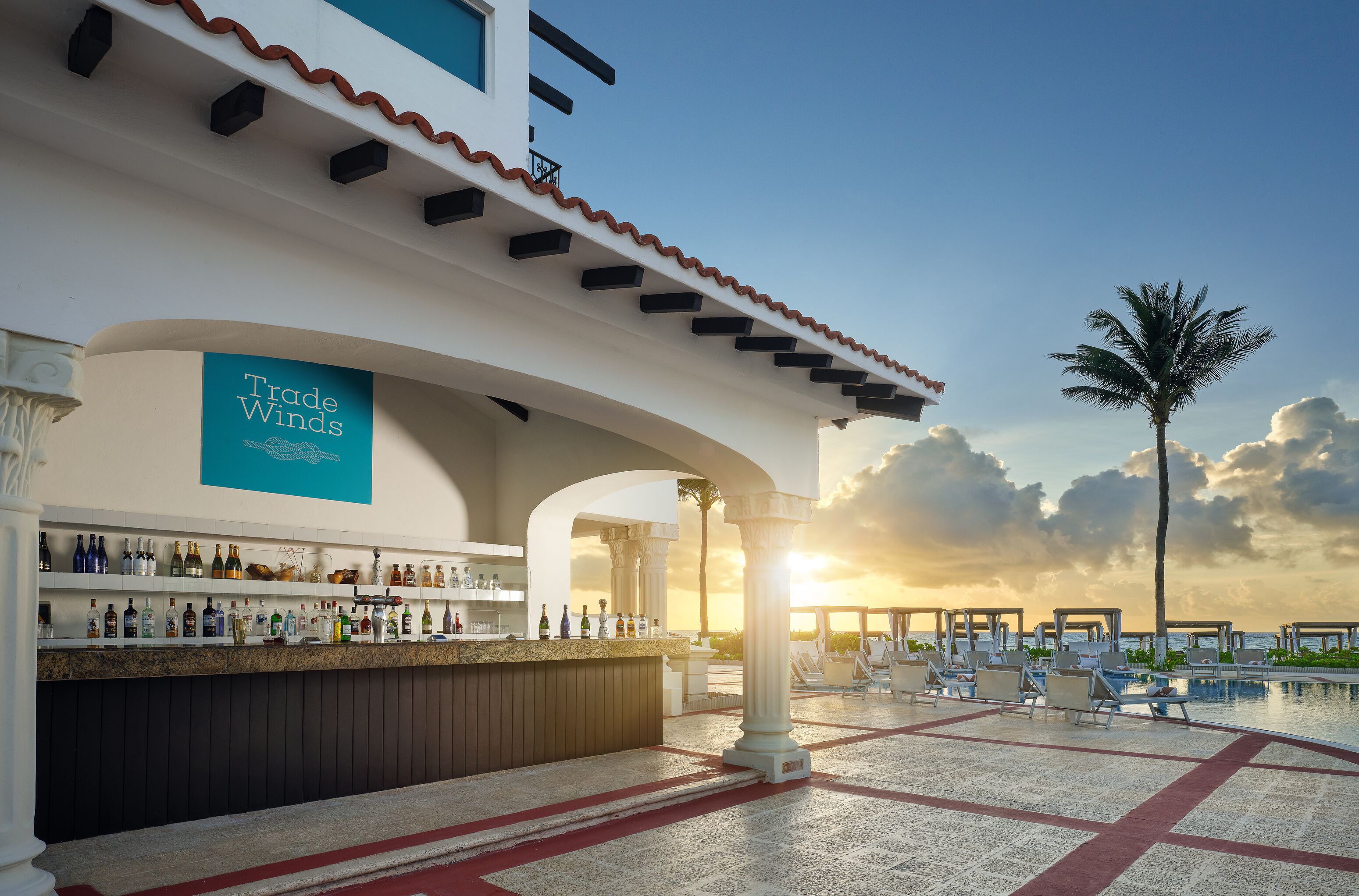 Zona de bar con un letrero 'Trade Winds', mesas al aire libre y piscina con vista al atardecer.