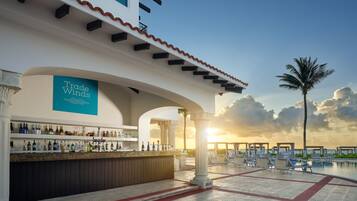 Zona de bar con un letrero 'Trade Winds', mesas al aire libre y piscina con vista al atardecer.