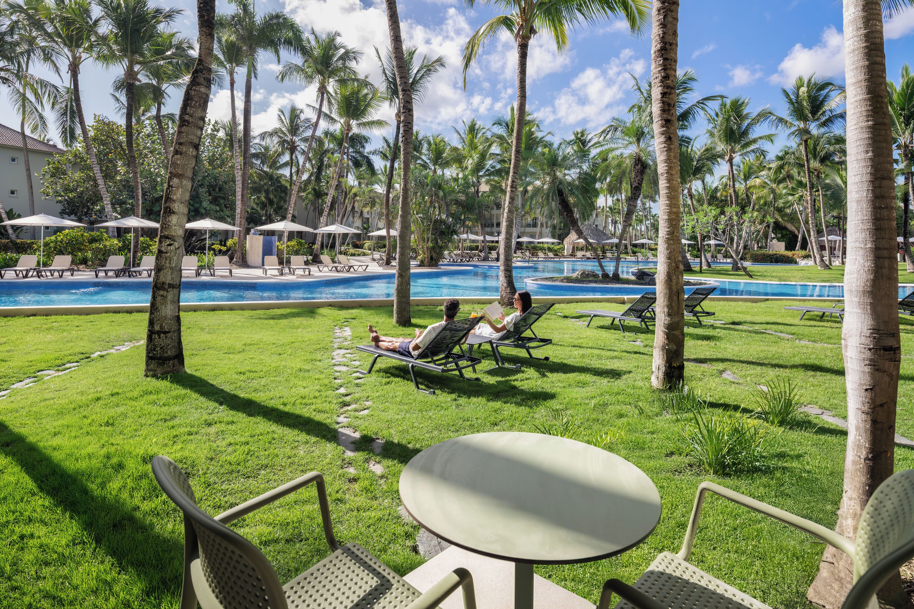 Zona de piscina con sillones de descanso y una mesa, rodeada de palmeras y un cielo despejado.