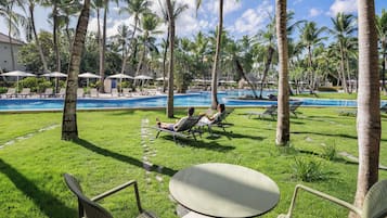 Zona de piscina con sillones de descanso y una mesa, rodeada de palmeras y un cielo despejado.