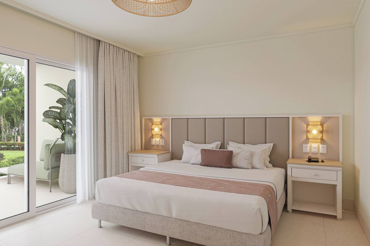 A modern bedroom with a large bed, two bedside tables with lamps, a sliding glass door to a balcony, and a potted plant.