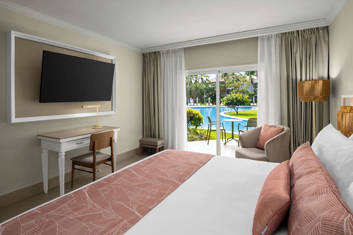 A hotel room with a bed, a desk, a chair, a TV, and a view of a pool and greenery outside.