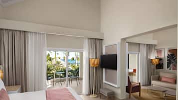 A hotel room with a bed, a TV, a sofa, a chair, a small table, and a balcony with a view of palm trees and a pool.