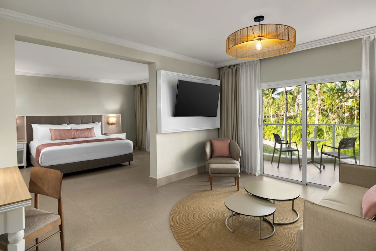 A hotel room with a bed, a TV, a seating area with a table and chairs, and a balcony with a view of palm trees.