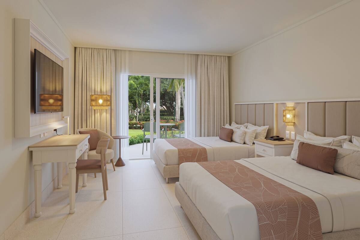 A hotel room with two beds, a desk, and a chair. There is a balcony with a view of greenery.