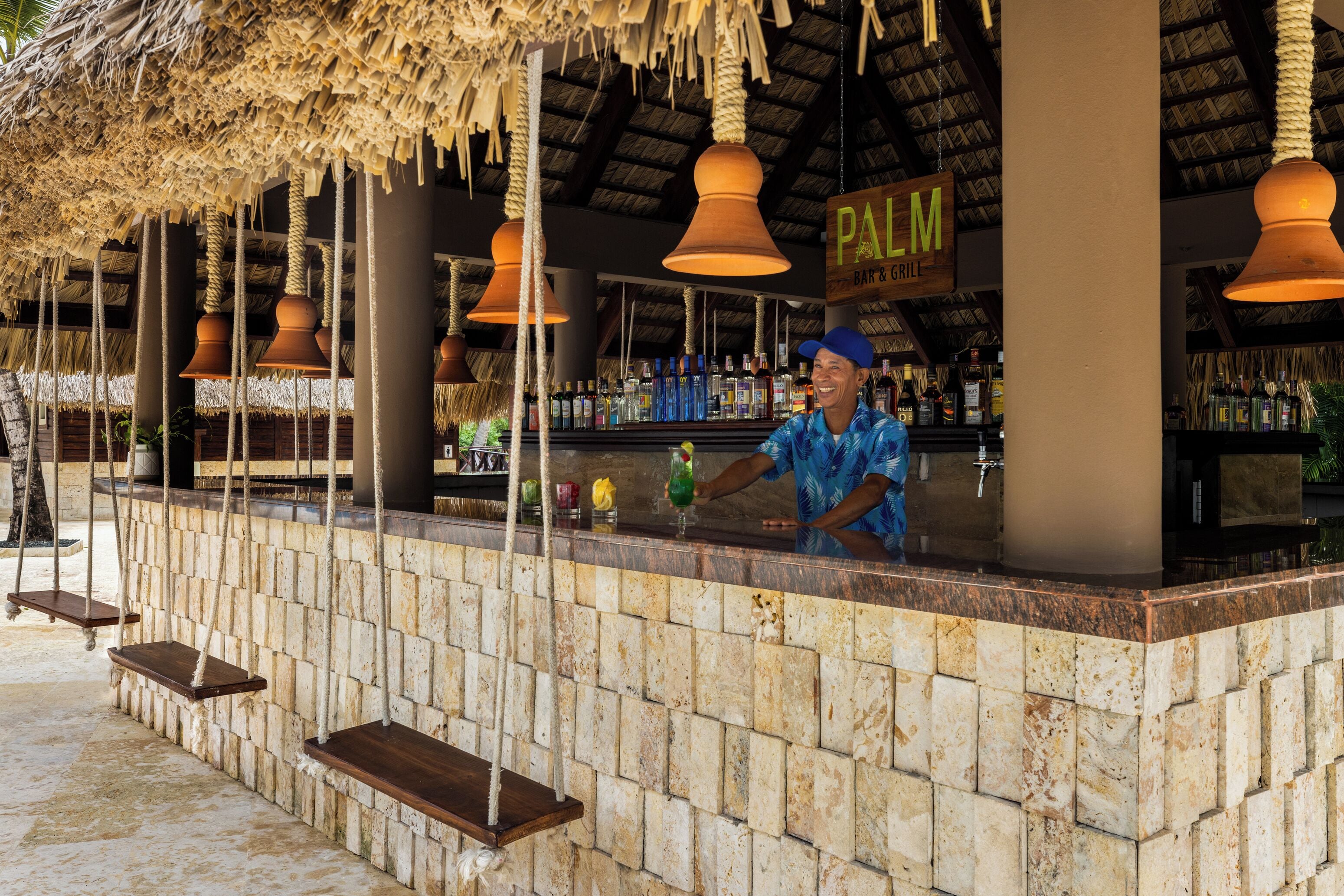 A bar area with a thatched roof, hanging pendant lights, and a sign reading "PALM BAR & GRILL".