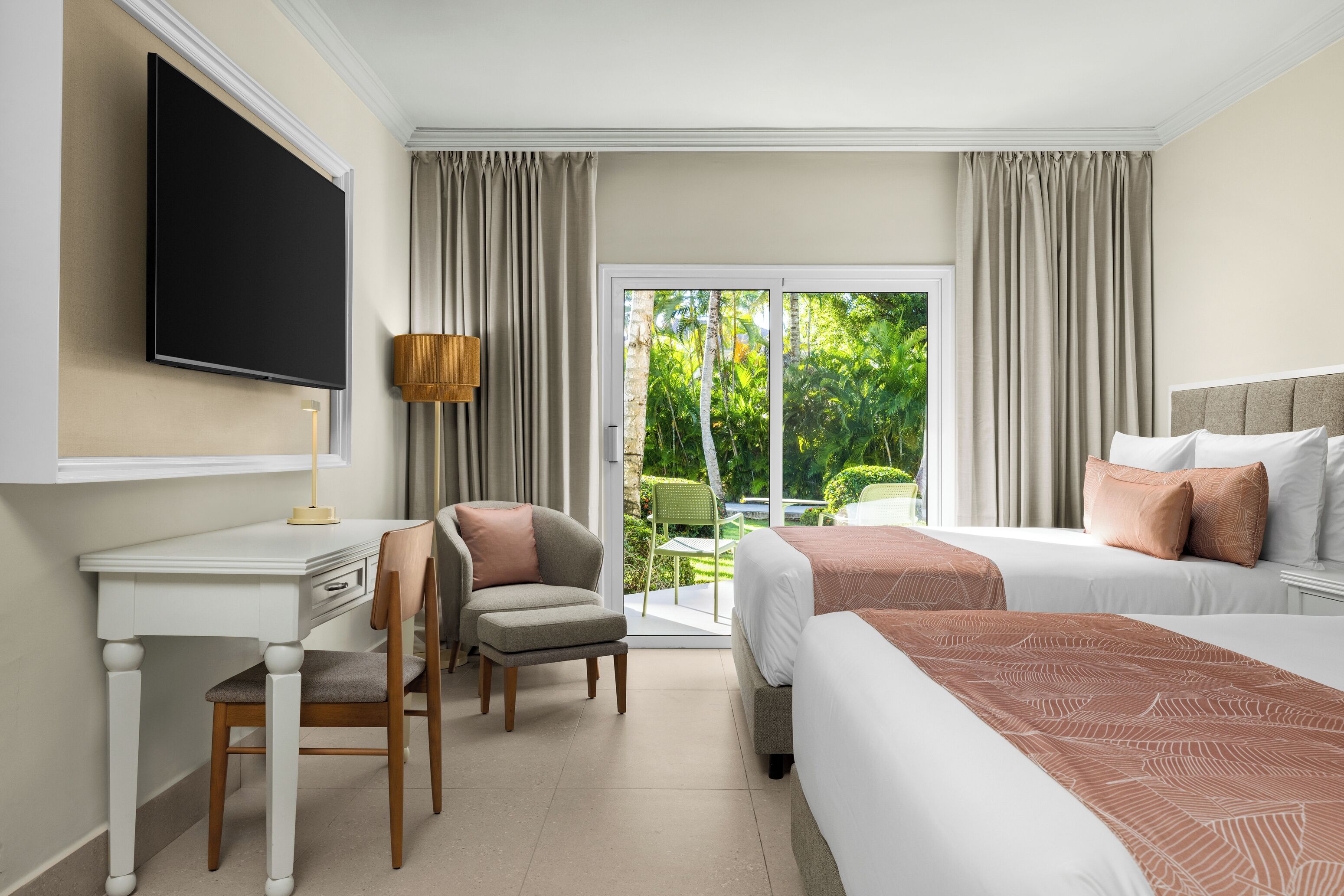 A hotel room with a flat-screen TV, a desk with a chair, two beds with pillows, a lamp, and a sliding glass door to a garden.