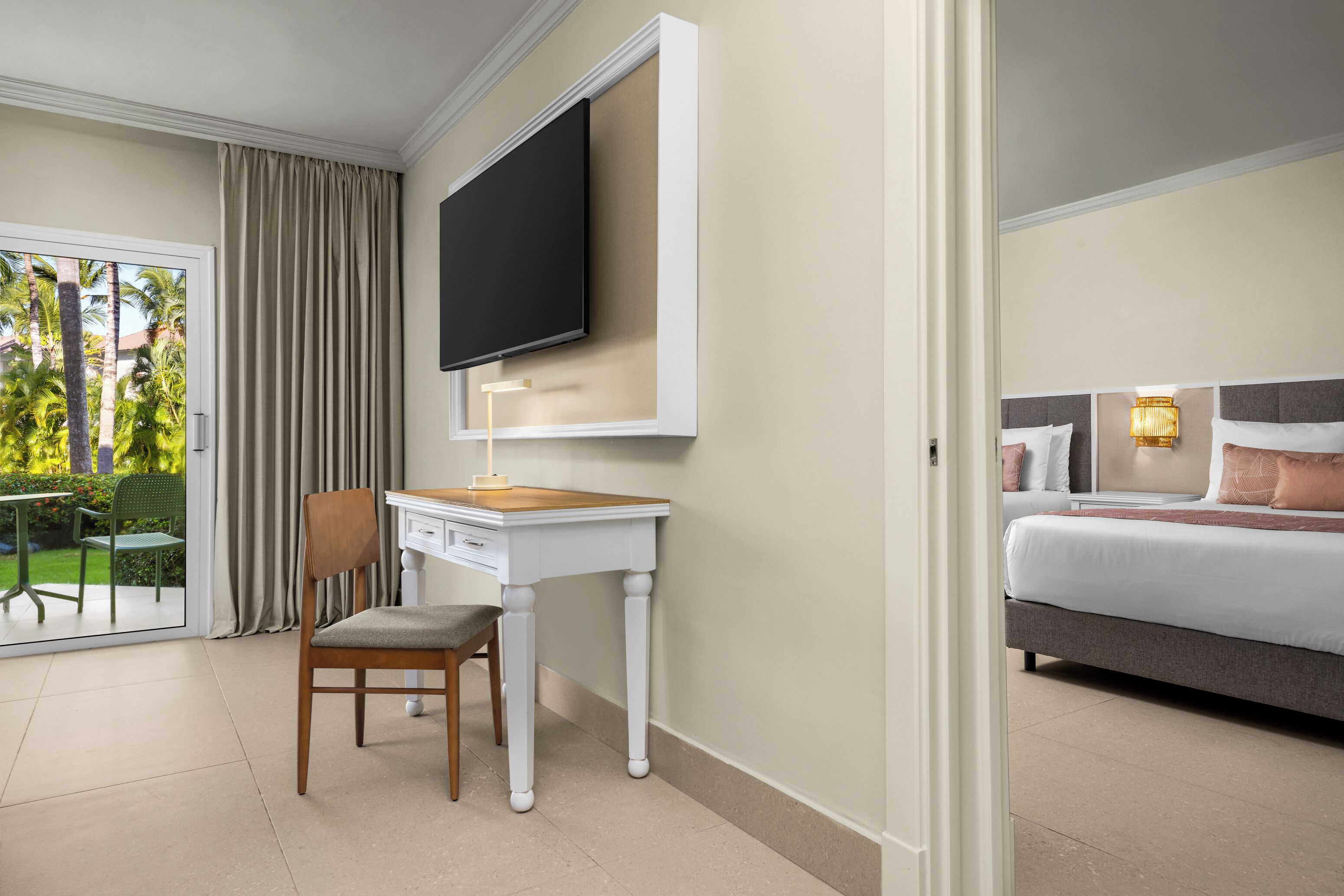 Een hotelkamer met een bed, een bureau met stoel, een aan de muur bevestigde televisie en uitzicht op een tuin door een glazen schuifdeur.