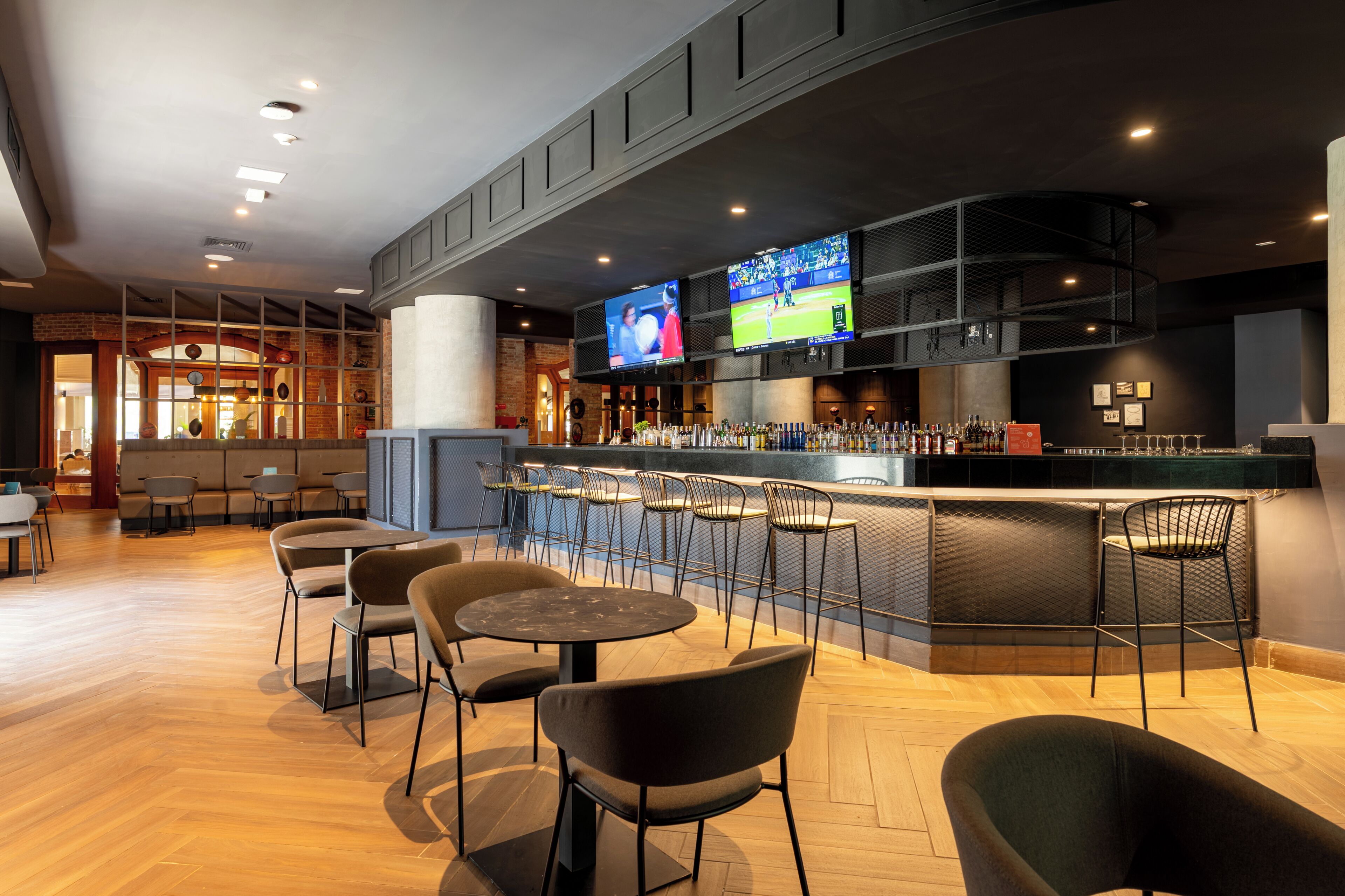 A bar area with multiple seating options, a counter with a beverage display, and two large screens mounted on the wall.