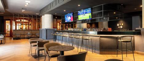 A bar area with multiple seating options, a counter with a beverage display, and two large screens mounted on the wall.