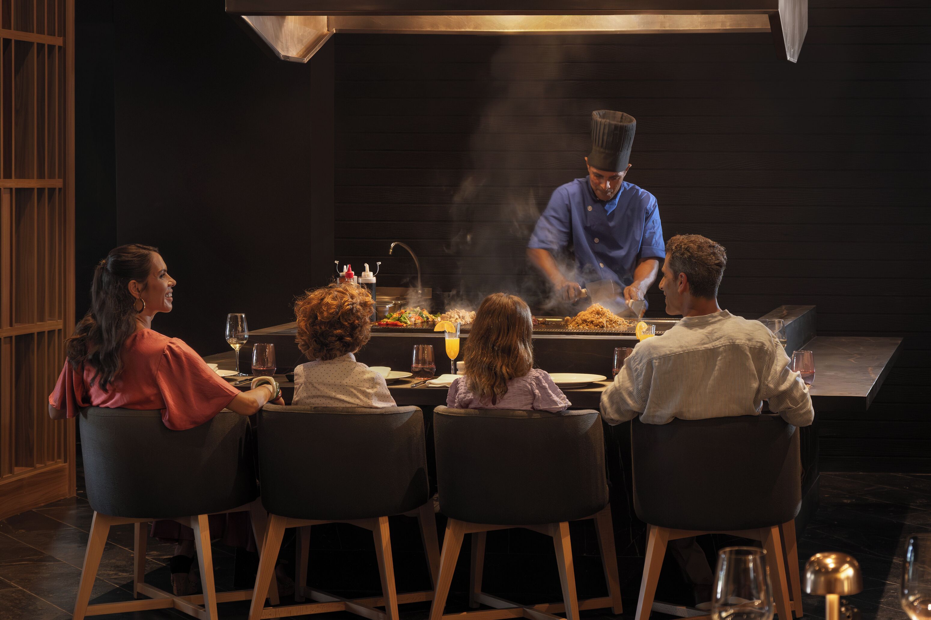 Un grupo de cuatro personas cenando en un restaurante, con un chef preparando comida al fondo.