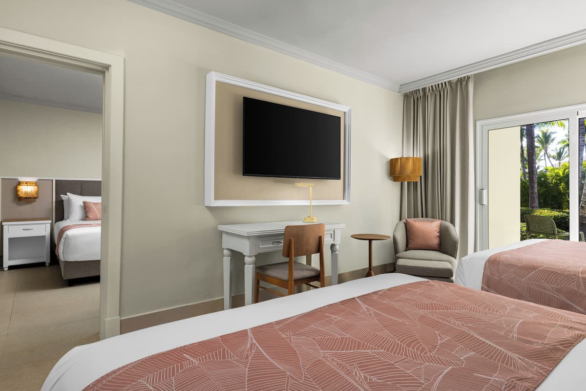 Ein Hotelzimmer mit zwei Betten, einem Schreibtisch, einem Stuhl, einem Fernseher und Blick auf Grünflächen.