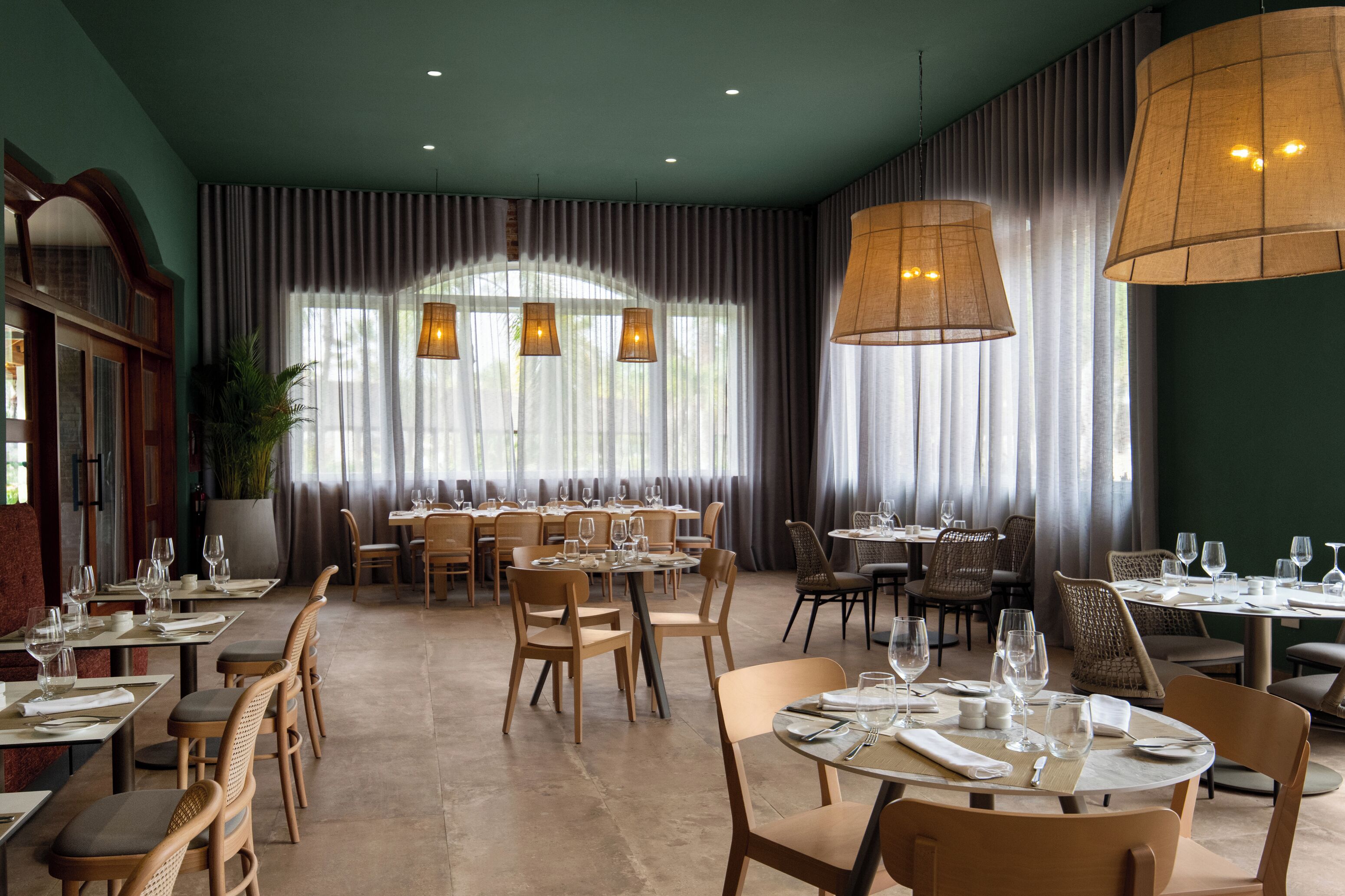 Un restaurante espacioso con techos altos, ventanas amplias con cortinas y varias mesas de comedor con sillas.