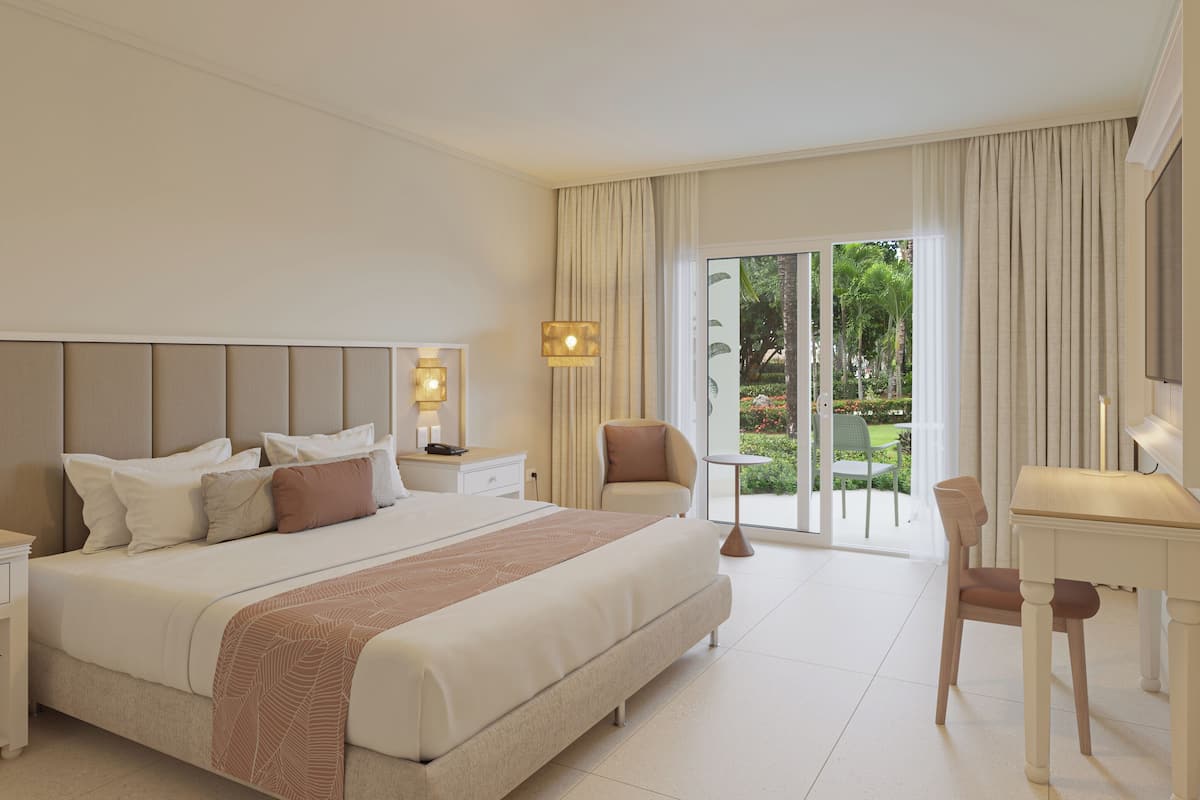 A hotel room with a large bed, a desk, and a chair. There is a view of a garden through the window.