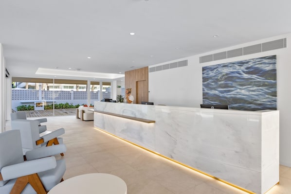 Un área de recepción moderna con mostrador de mármol, una obra de arte grande que representa agua y vistas a una piscina y asientos exteriores.