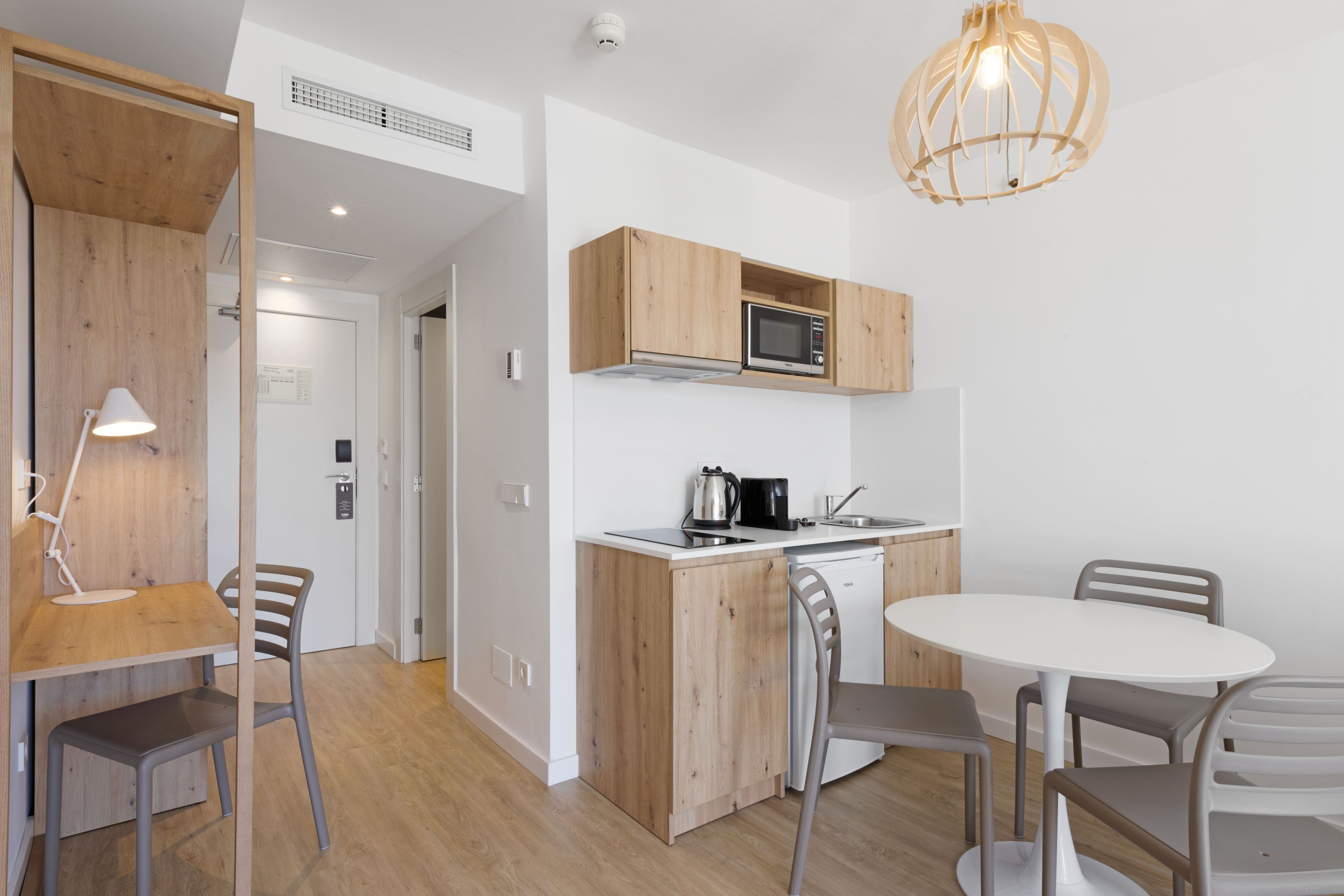 Cocina compacta con armarios de madera, microondas, hervidor y un comedor con sillas y mesa.