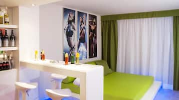 Una habitación de hotel moderna con cama, escritorio con taburetes, minibar y una obra de arte en la pared.