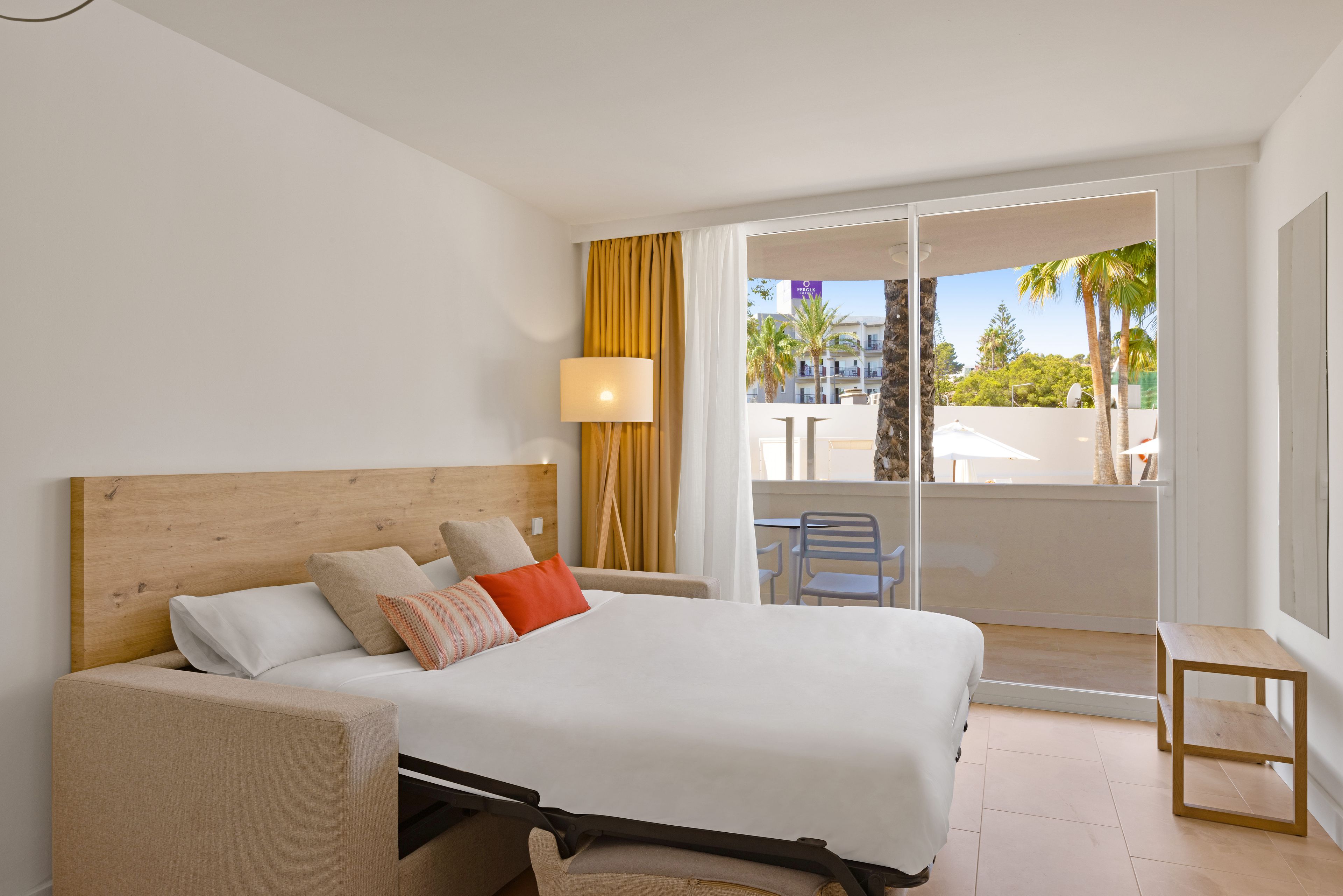 Habitación de hotel con cama, mesita de noche, lámpara y vistas al exterior.