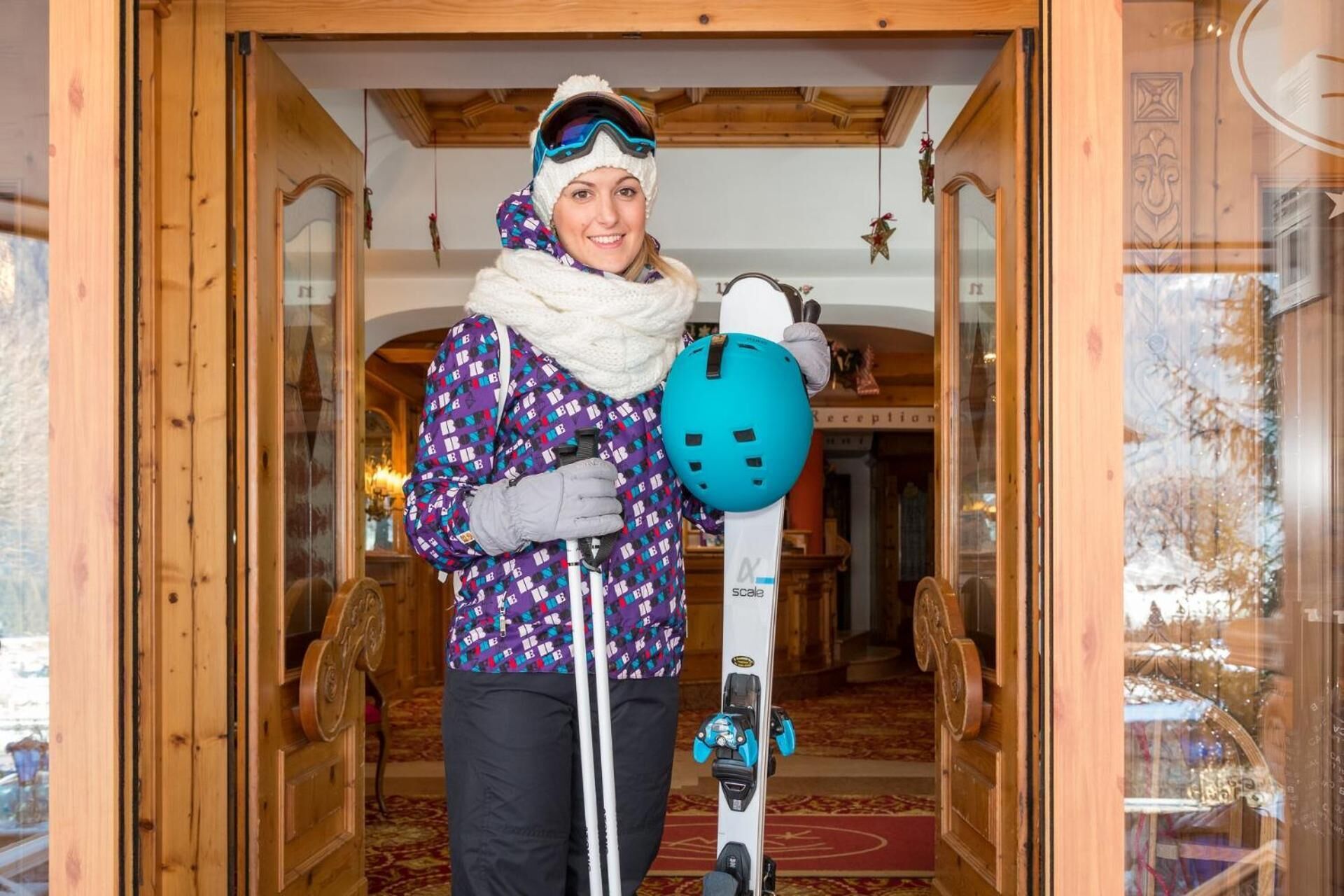 A person holding skis and a helmet in a wooden interior with a reception area visible in the background.