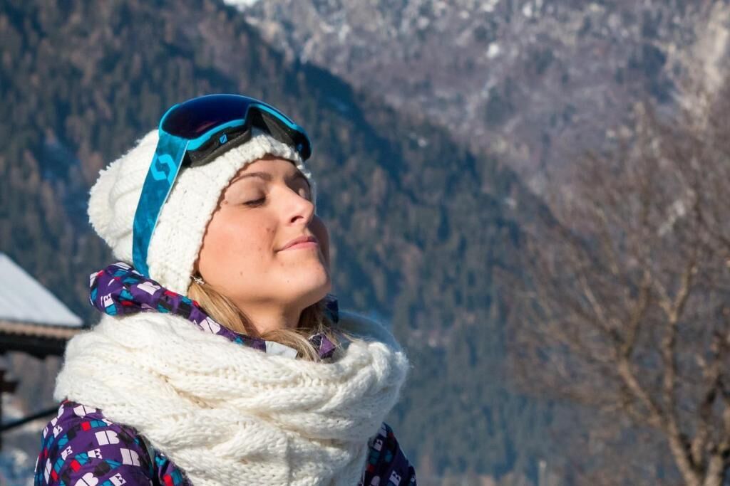 Una persona con gorro y bufanda, de pie al aire libre con montañas al fondo.