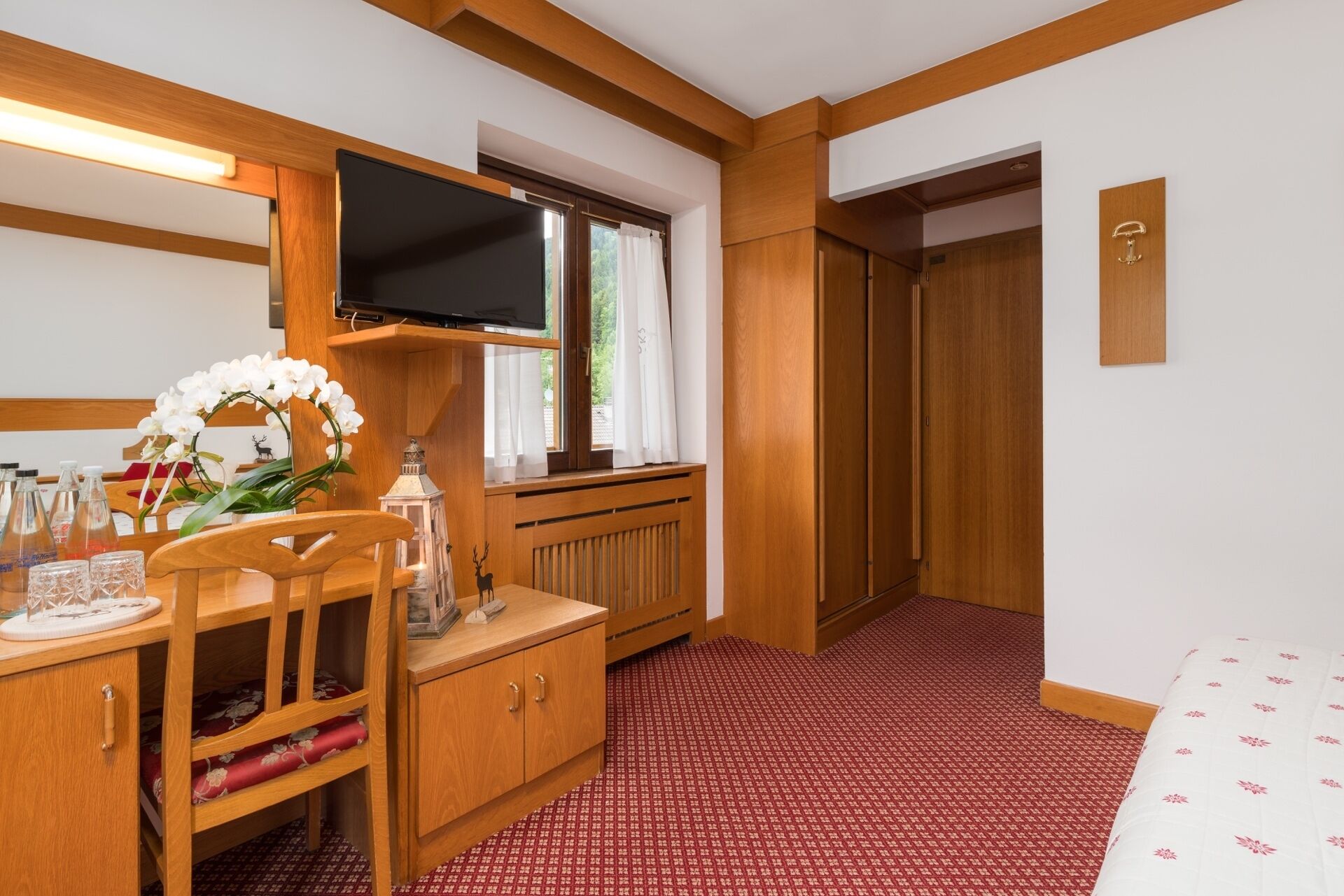 Ein Hotelzimmer mit einem Holztisch, einem an der Wand befestigten Fernseher, einem Bett mit einem gemusterten Bettüberwurf und einem roten Teppichboden.