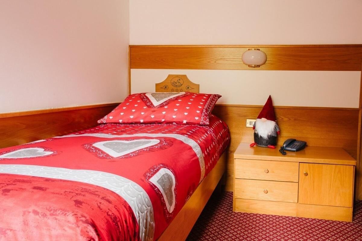 A neatly made bed with a red and white patterned bedspread and pillows, a wooden nightstand with a small gnome figurine and a phone, and a simple wooden headboard.