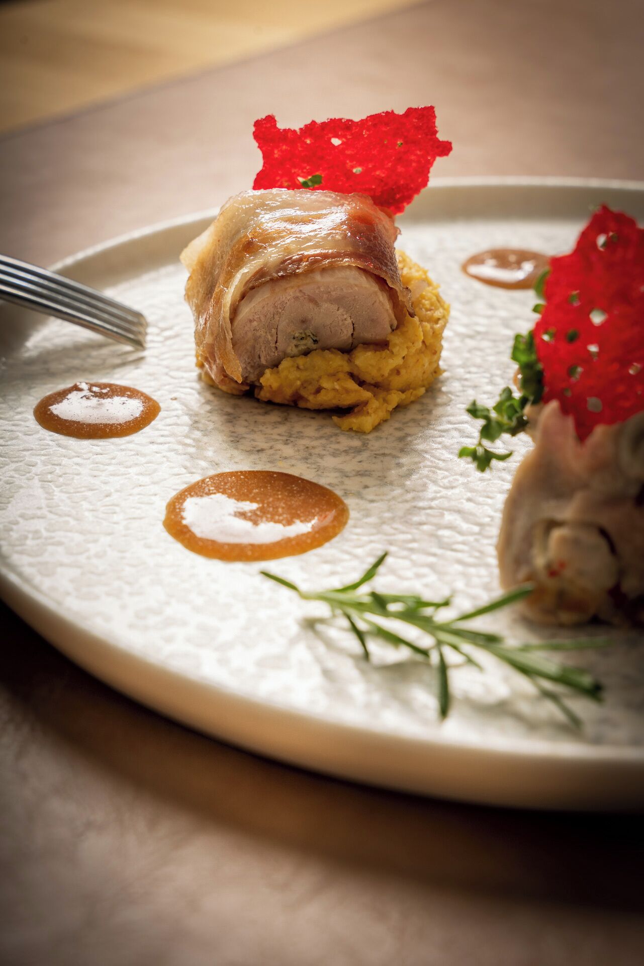 A plated dish with a rolled meat, a red garnish, and a yellow puree, served on a textured white plate.