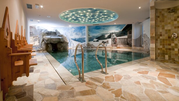 An indoor swimming pool with a stone floor, wooden benches, and a mural of a mountainous landscape. - Hotel Gianna (Tre Ville)