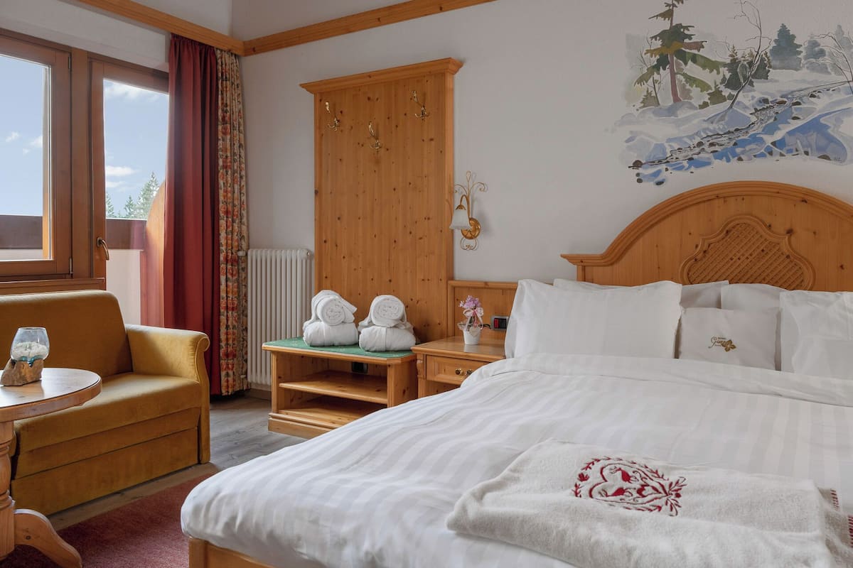 A hotel room with a bed, a wooden wardrobe, a chair, a table, and a painting on the wall.