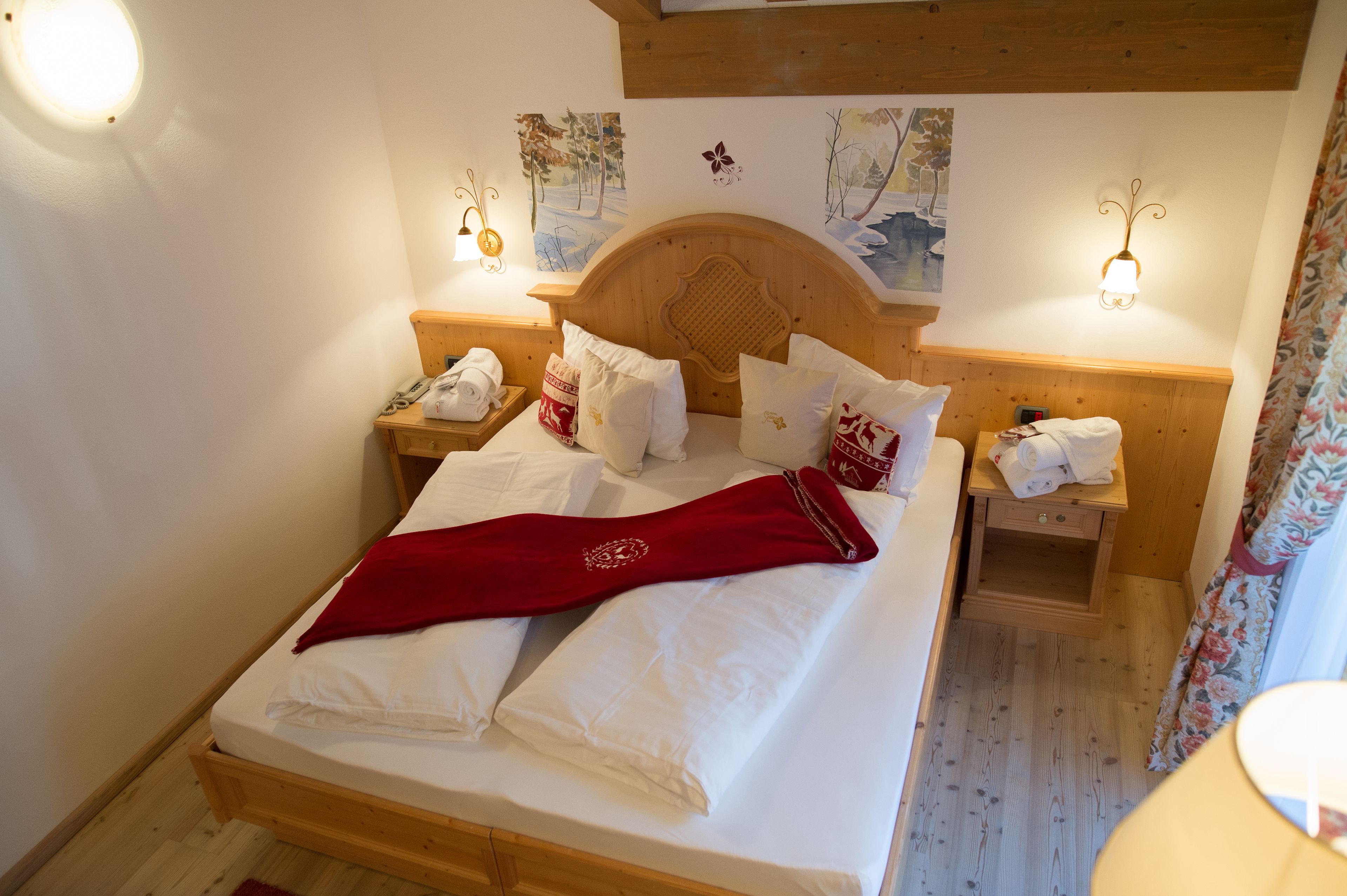 Ein ordentlich bezogenes Bett mit weißen Leinen und einer roten Decke, ein h ölzernes Kopfteil, zwei Nachttische mit Lampen sowie Wandbilder mit Winterlandschaften.