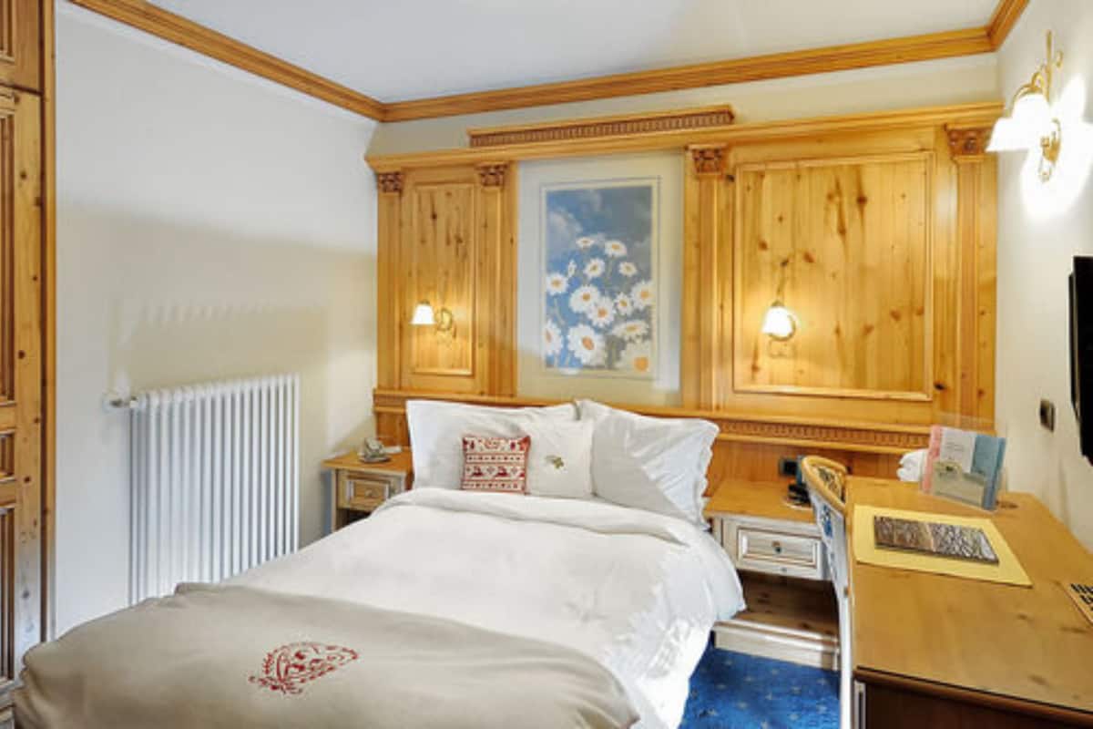 A hotel room with a wooden headboard, a bed with white linens, a nightstand with a lamp, a desk with a chair, and a framed floral painting on the wall.