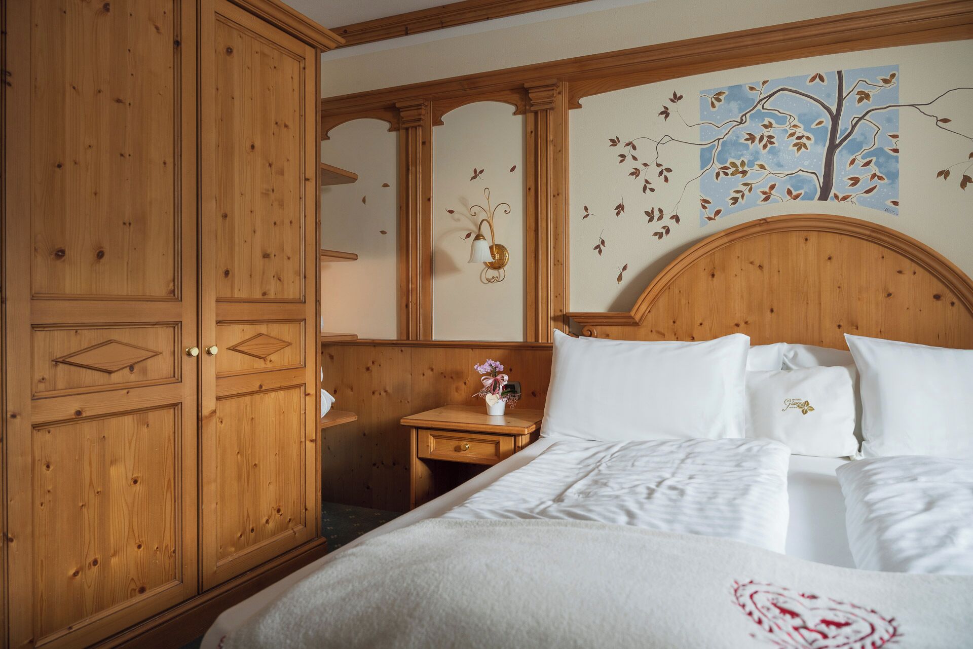A bedroom with a wooden wardrobe, a nightstand with a vase, and a bed with white bedding.