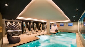 Un área de piscina interior moderna con sillones de descanso, un jacuzzi y una pared de piedra.