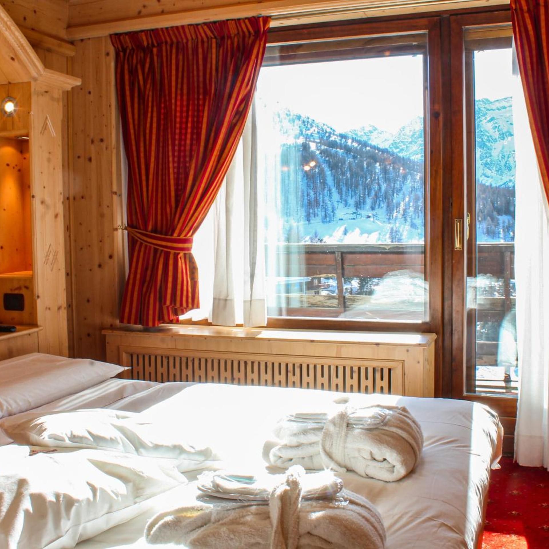 A wooden cabin room with a bed, a window with curtains, and a view of a snowy landscape.
