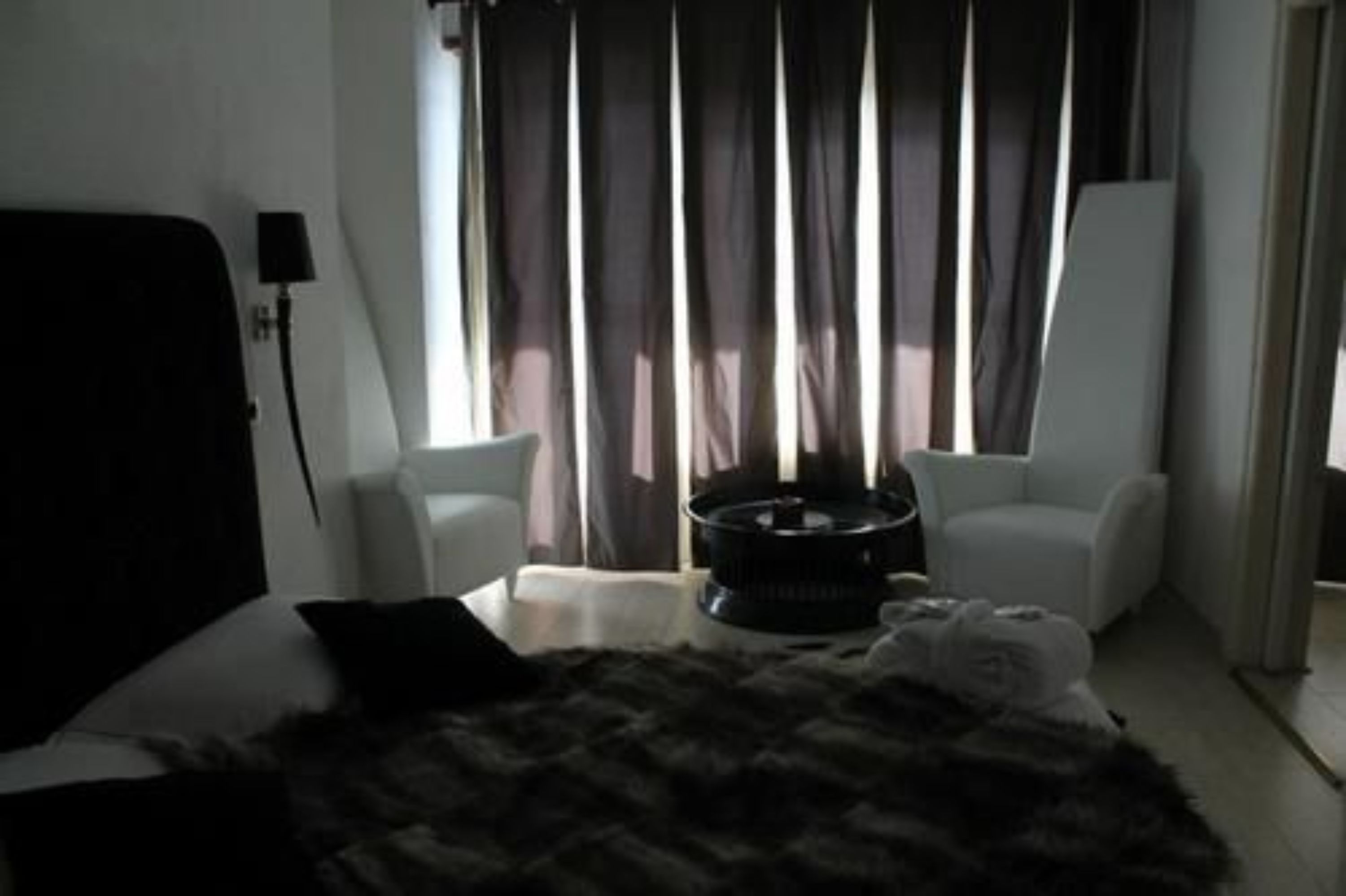 A modern bedroom with a large bed, a black furry throw, two white armchairs, and a black speaker.