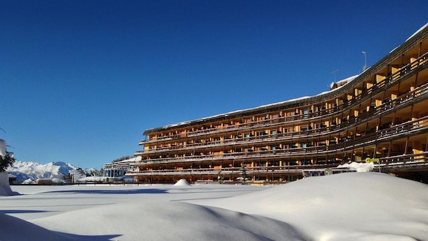 Een hotel met meerdere verdiepingen, balkons, een met sneeuw bedekte grond en een helderblauwe lucht.
