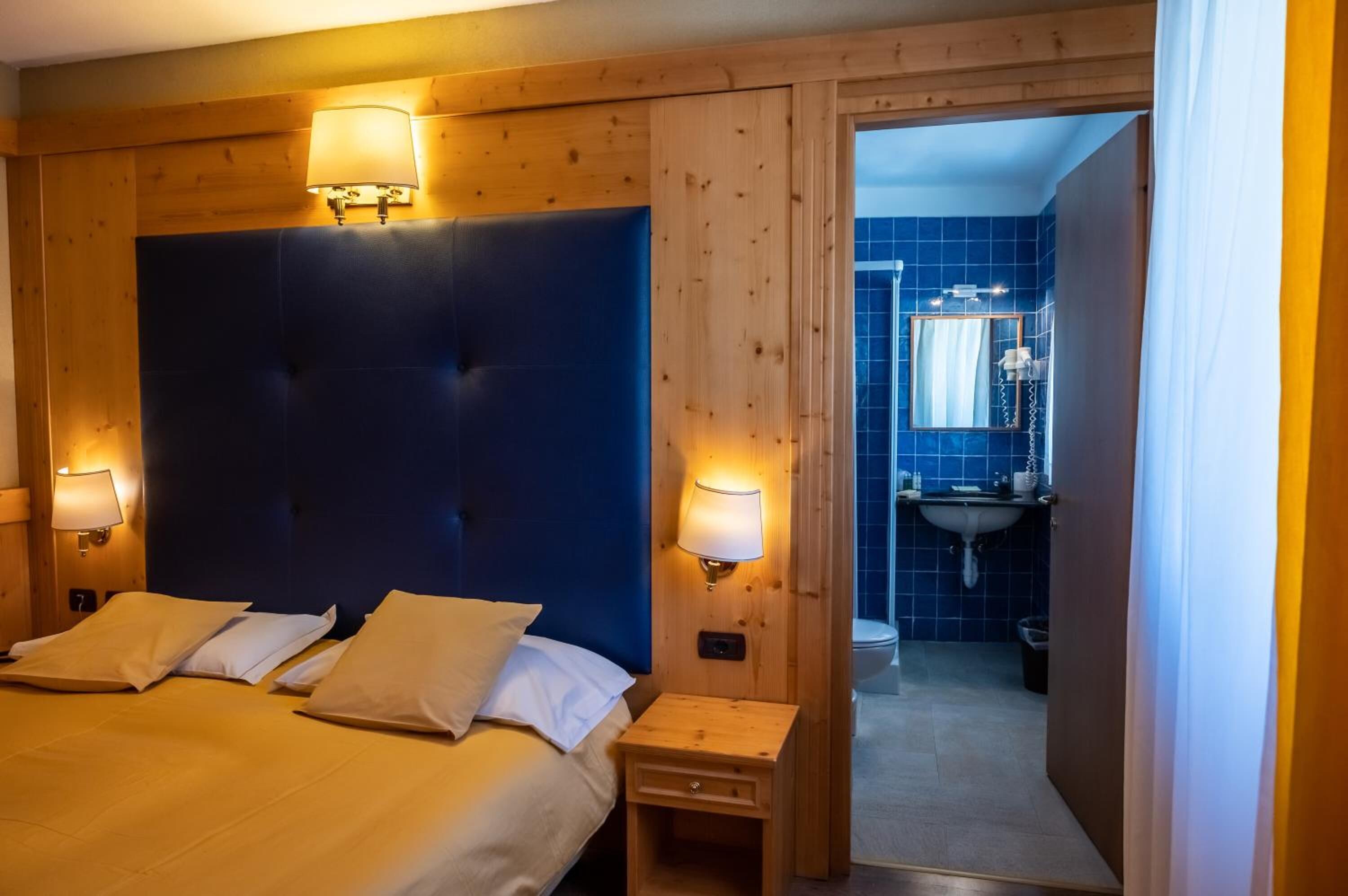 A hotel room with a large bed, two bedside lamps, a wooden headboard, a small bedside table, and a bathroom visible through an open door.
