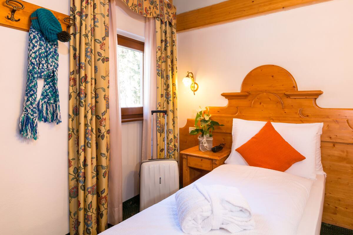 A hotel room with a wooden headboard, a bedside table with a lamp, a window with floral curtains, and a towel on the bed.
