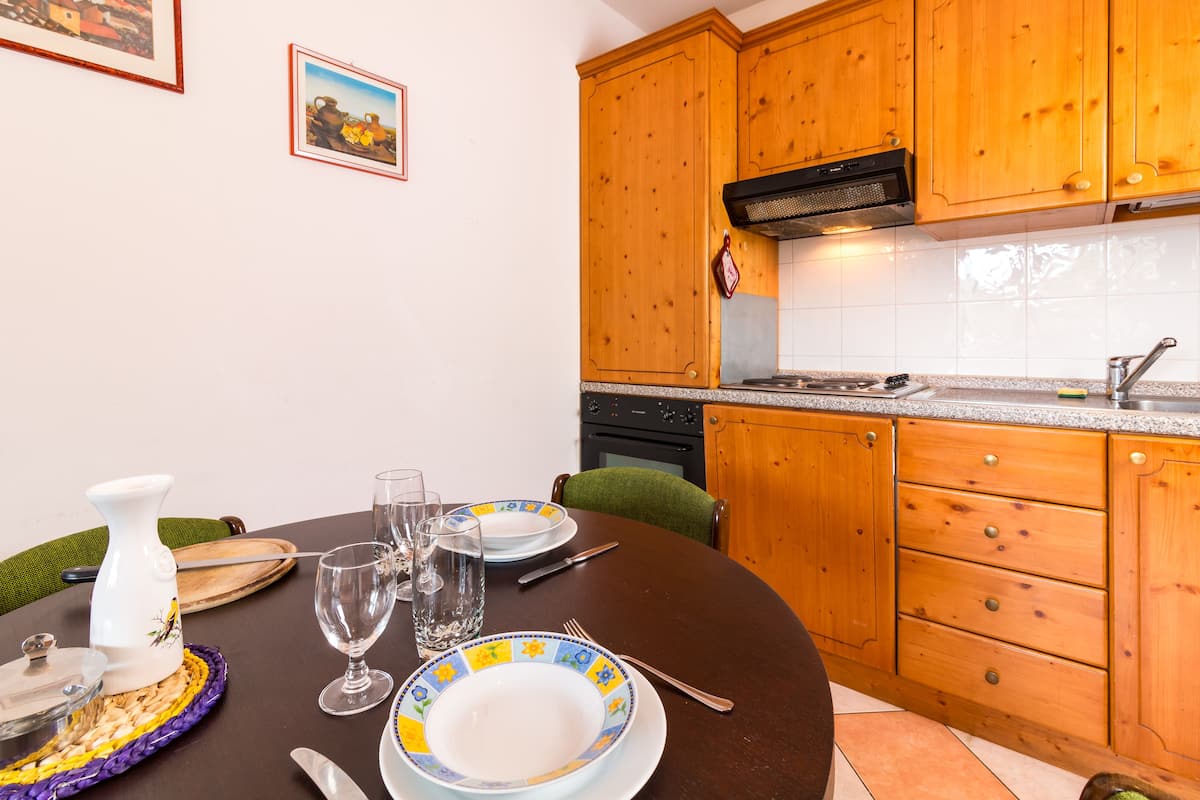 A dining area with a round table set for four, including plates, glasses, and cutlery. Wooden cabinets and a tiled backsplash are visible in the background.