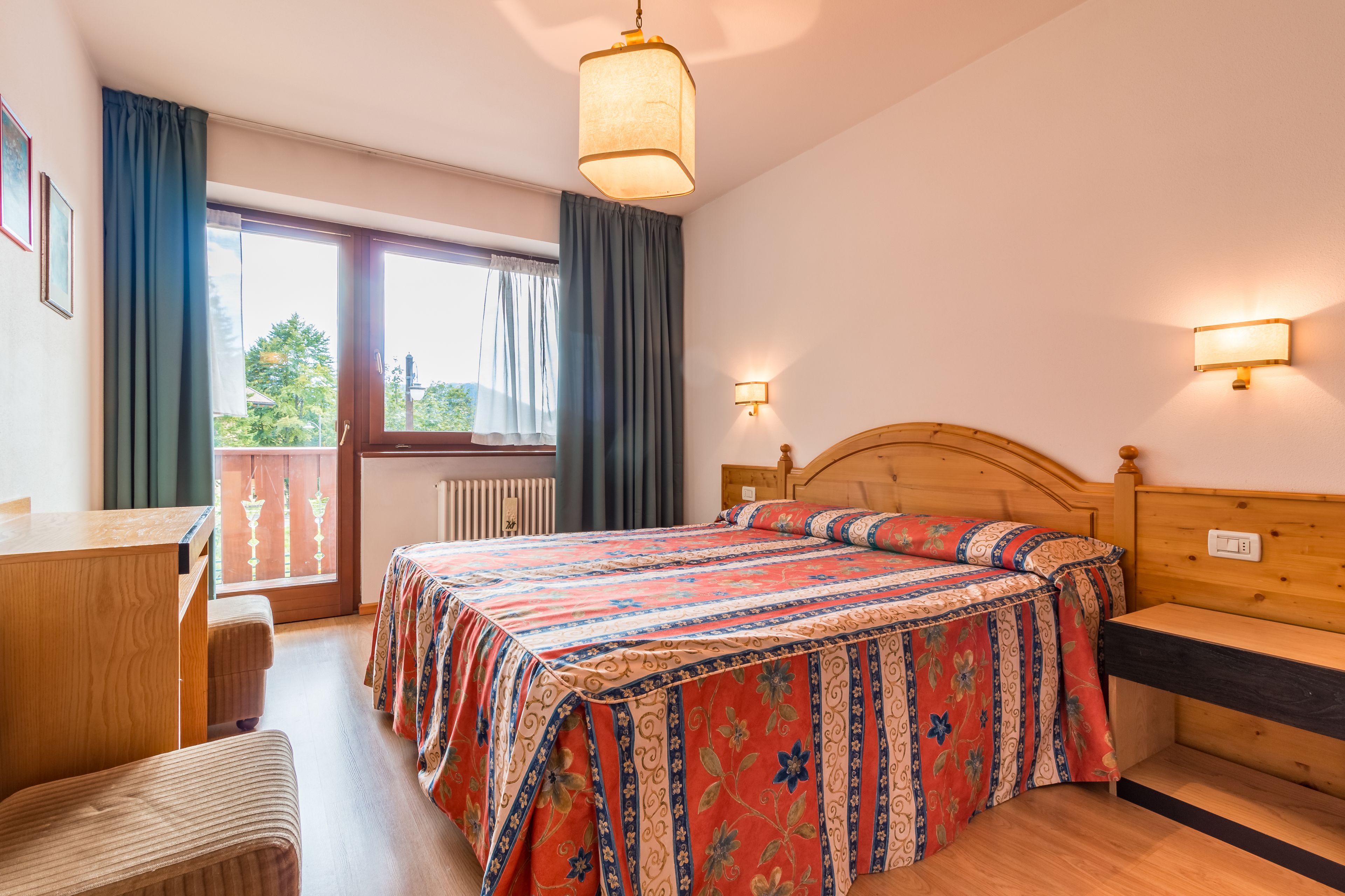 Ein Hotelzimmer mit Bett, Schreibtisch, Stuhl, einem Fenster mit Vorhängen und Blick ins Grüne.
