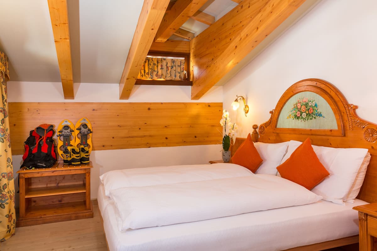 A bedroom with a wooden ceiling, a bed with white bedding and orange pillows, a nightstand with a lamp, and a window with a view of the sky.
