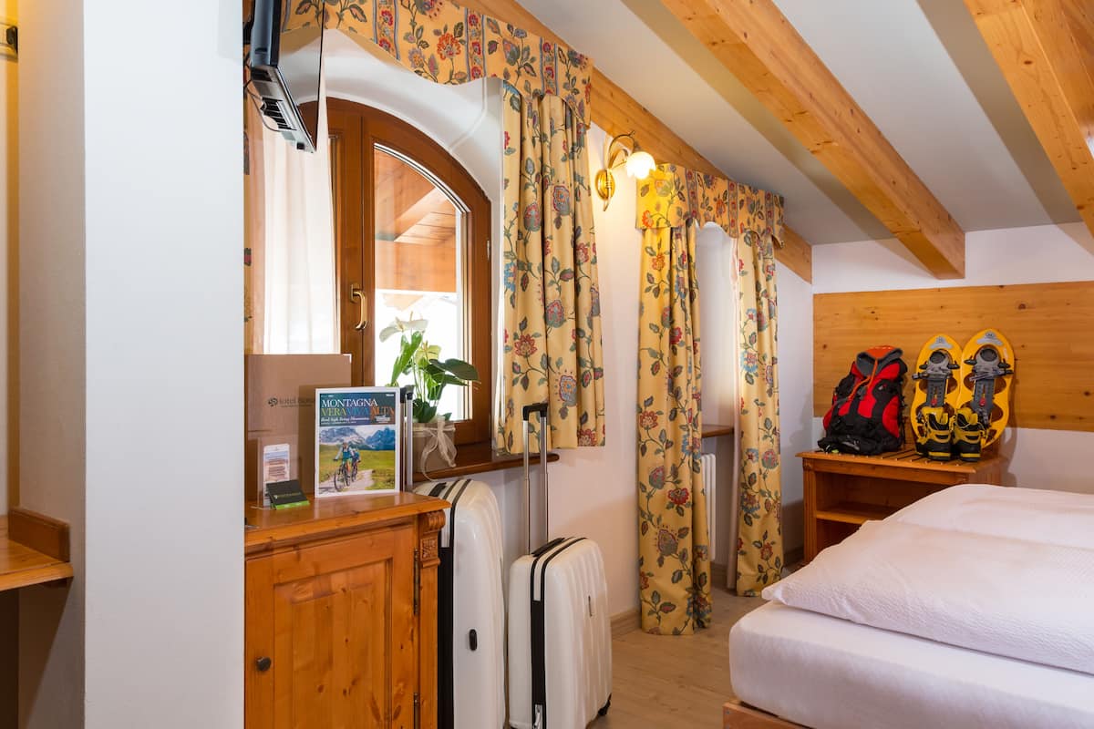 A hotel room with wooden beams, a bed, a desk with a suitcase, a mirror, and a window with floral curtains.