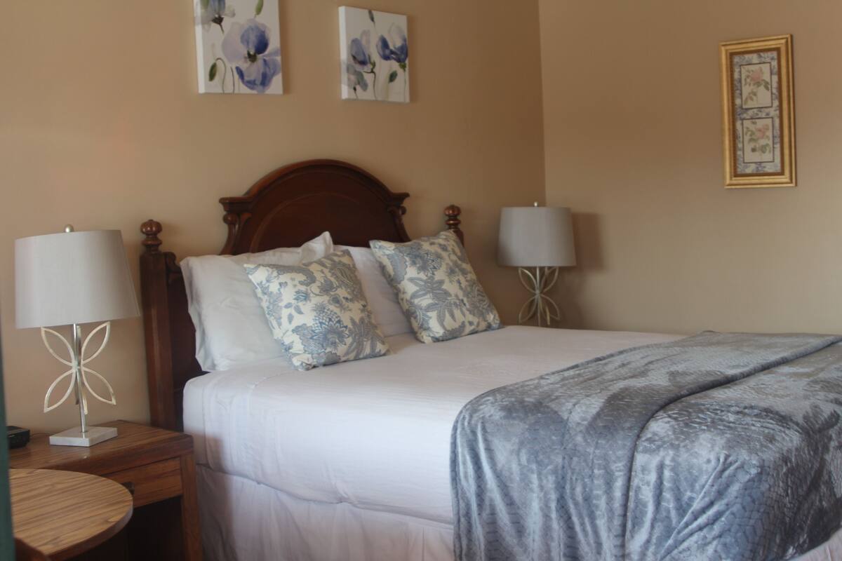 A neatly made bed with a grey blanket, two matching lamps on either side, and floral artwork on the walls.